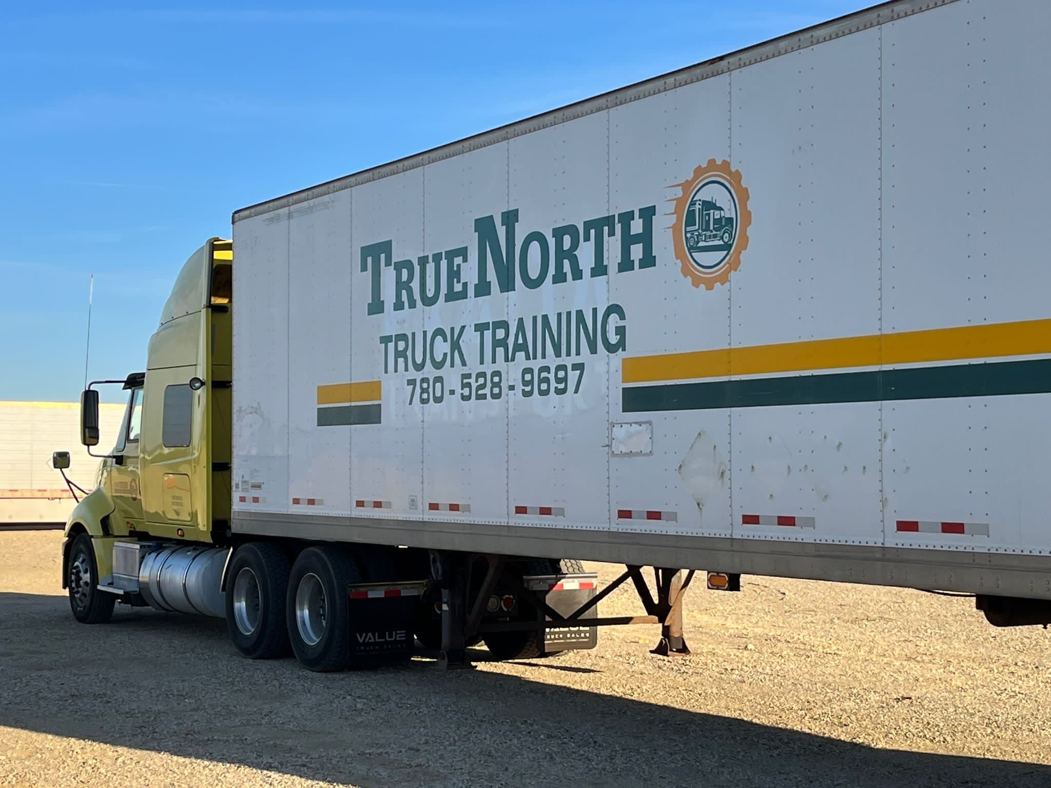 Side view of a True North Truck Training semi-truck and white trailer parked in a gravel lot